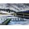 An icy, frozen river with a deck extends onto it. A bridge on right crosses the other side, a riverbank of Winter grasses with a snowy hill.